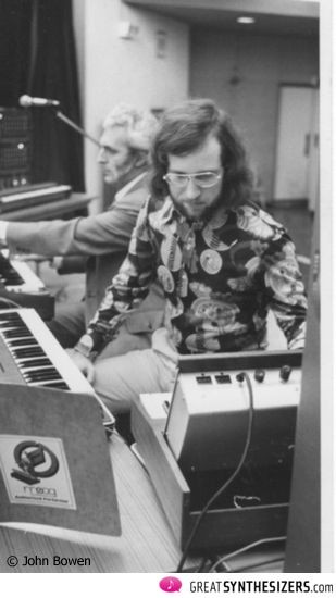 Two men play electronic keyboards and synthesizers in a music studio. The man in the foreground has glasses and a patterned shirt, while the man in the background appears focused on another keyboard. Black and white photo.