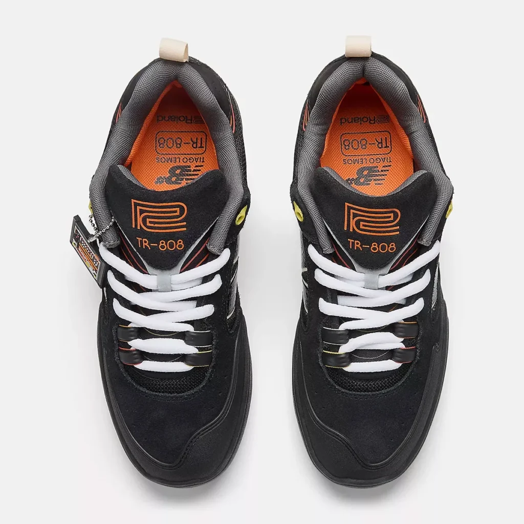 Top view of a pair of black sneakers with white laces, orange insoles, and TR-808 branding on the tongues, featuring yellow accents and Roland logos.