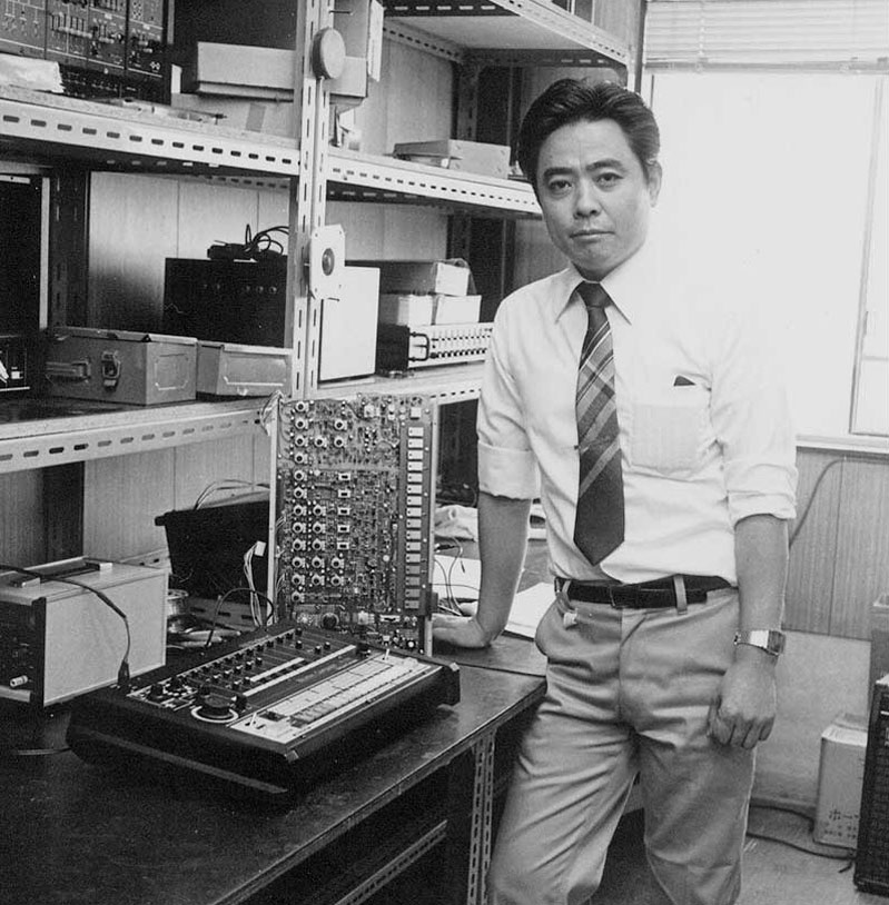 A man in a dress shirt and tie stands in an electronics lab, resting his hand on a table with audio equipment and circuit boards. Shelves with electronic devices and components line the walls behind him.