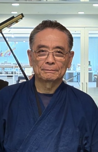 An older man with short gray hair, glasses, and a blue robe stands indoors, smiling softly. There is a piano and glass doors in the background, with a bright, modern room visible through the glass.