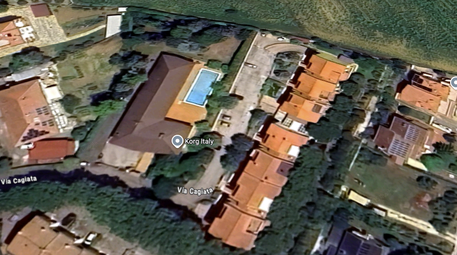 A satellite view of a residential area in Italy, showing an L-shaped building with a pool, several orange-roofed houses, parked cars, trees, and green fields nearby. Korg Italy and Via Cagliari are marked on the image.