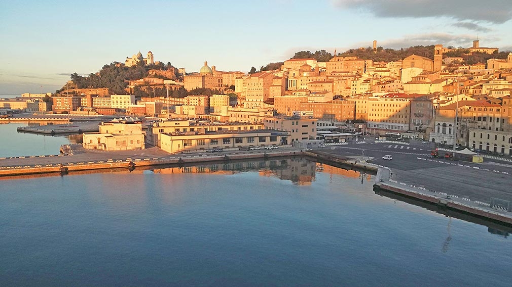 A coastal city with historic buildings bathed in warm sunlight, perched on a hill beside a calm harbor with piers and waterfront structures. The sky is clear with a few clouds.