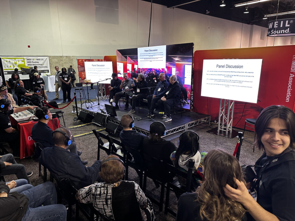 A group of people attend a panel discussion at an indoor event. Panelists sit onstage with microphones, and two large screens display Panel Discussion. Audience members wear headphones and face the stage.