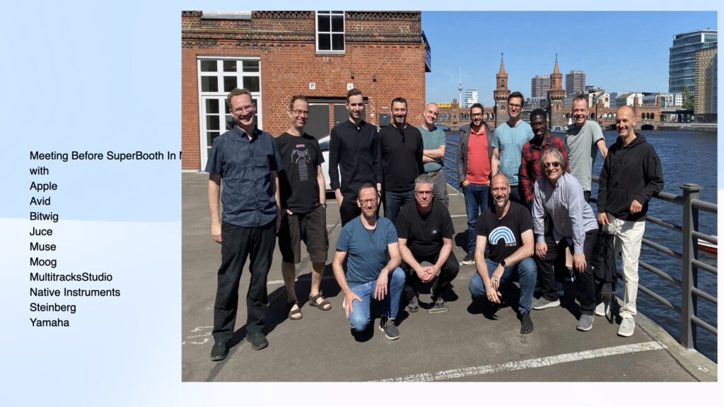 A group of 15 people pose and smile outdoors by a riverside railing, with brick buildings and the Oberbaum Bridge in the background. A light blue box lists music tech brands like Apple and Steinberg on the left side.