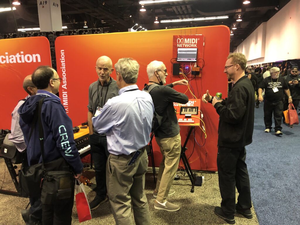 A group of people gather around a booth displaying vintage synthesizers and posters at a convention. One person plays an organ while others observe, and various banners and promotional materials are visible.