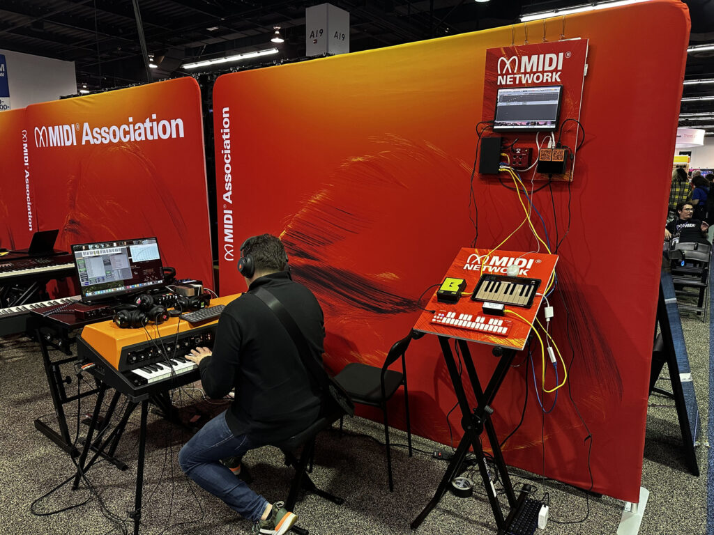 A person sits at a keyboard workstation with music equipment and a laptop, next to a bright red display with MIDI Association branding and MIDI devices mounted on a stand and wall.