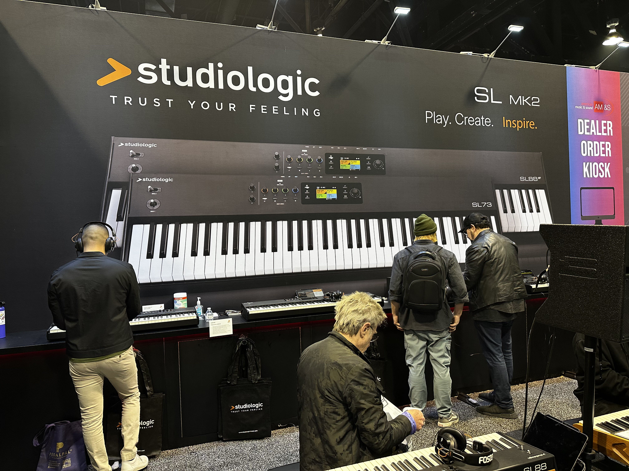 Three people examine keyboards at a Studiologic booth displaying SL MK2 digital pianos. A large banner with the Studiologic logo and slogan is behind them, and smaller keyboards are arranged on tables beneath the banner.
