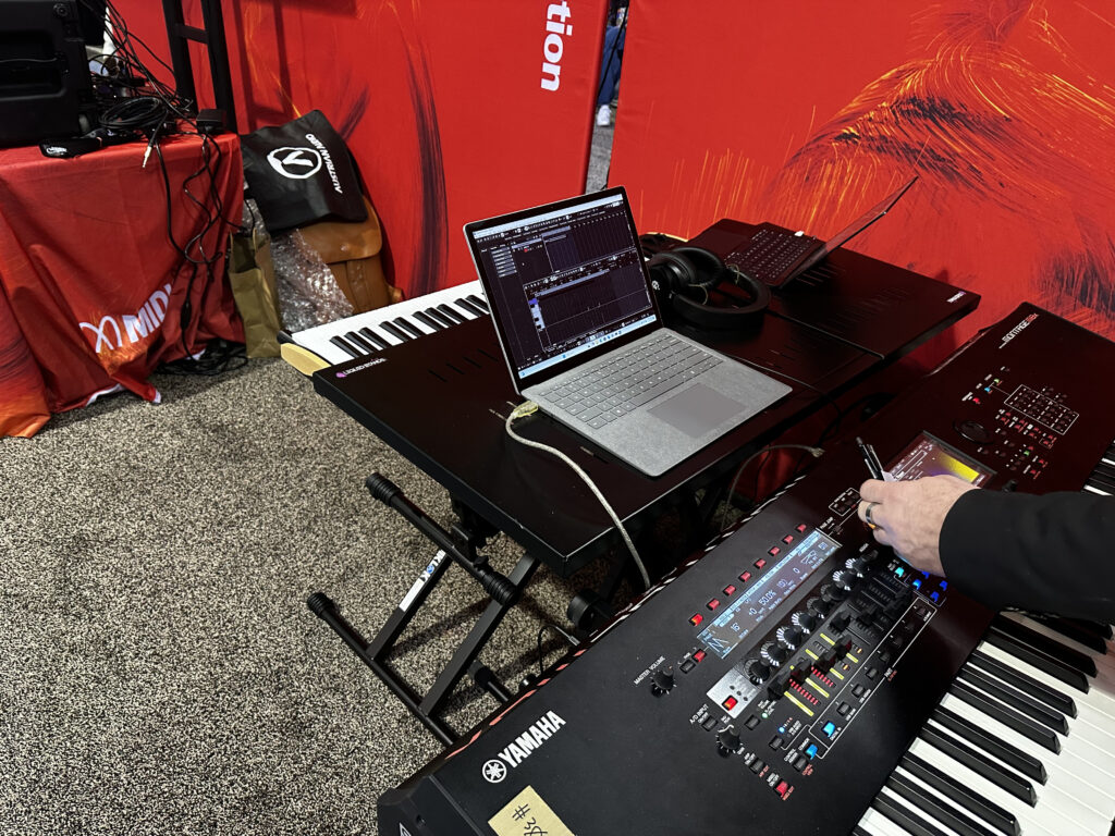 A close-up of a music production setup with two electronic keyboards, a laptop displaying audio software, headphones, and a person adjusting settings on a Yamaha keyboard. Red banners and carpeted flooring are visible in the background.