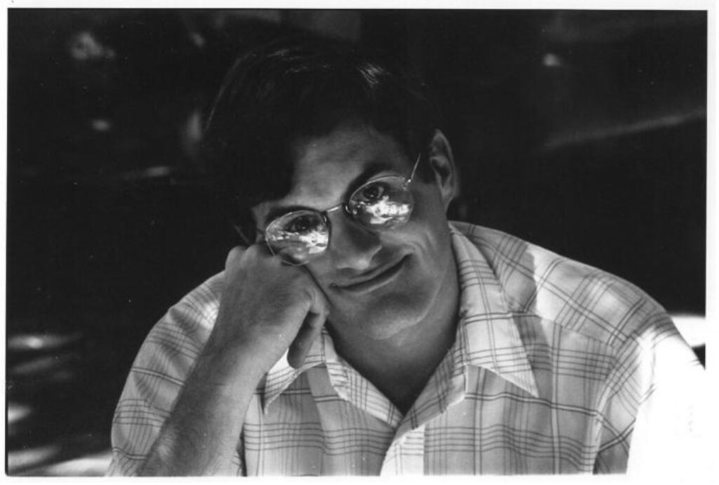 Black-and-white photo of a man with dark hair wearing glasses and a plaid shirt, resting his face on his hand and smiling slightly. Reflections are visible in his glasses.