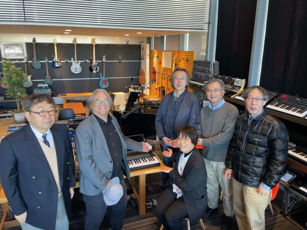 Six people pose in a music studio filled with keyboards, synthesizers, and guitars on the wall; most are standing, with one person seated and smiling near a keyboard.