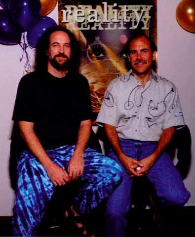 Two men sit side by side in front of a poster that says reality. The man on the left has curly hair, a beard, and wears a black shirt and blue tie-dye pants. The man on the right has a mustache and wears a patterned shirt.