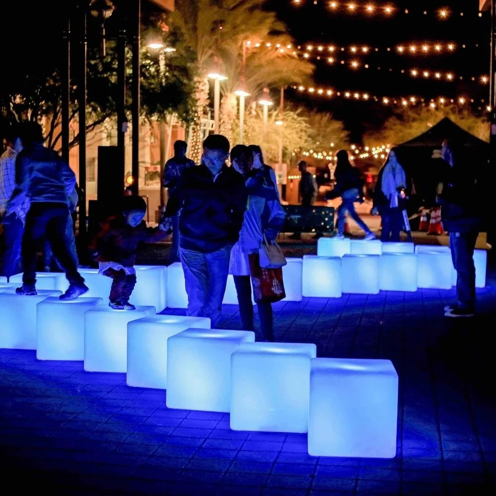 People walk and play on glowing blue cubes at night, surrounded by string lights and illuminated trees in an outdoor urban setting. The scene has a festive, lively atmosphere.