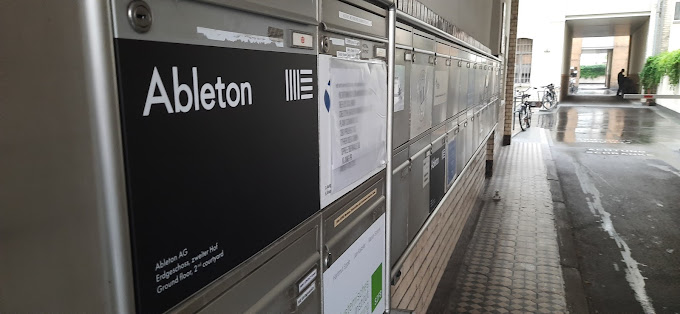 A row of mailboxes in a covered outdoor corridor, with one labeled Ableton in large letters. The ground is wet, bicycles are visible in the background, and plants hang on the wall at the end of the corridor.