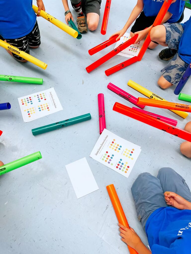 Children sitting in a circle on a light floor, each holding colorful plastic tubes (Boomwhackers), with music sheets featuring colored dots in the center. Only their legs and hands are visible.