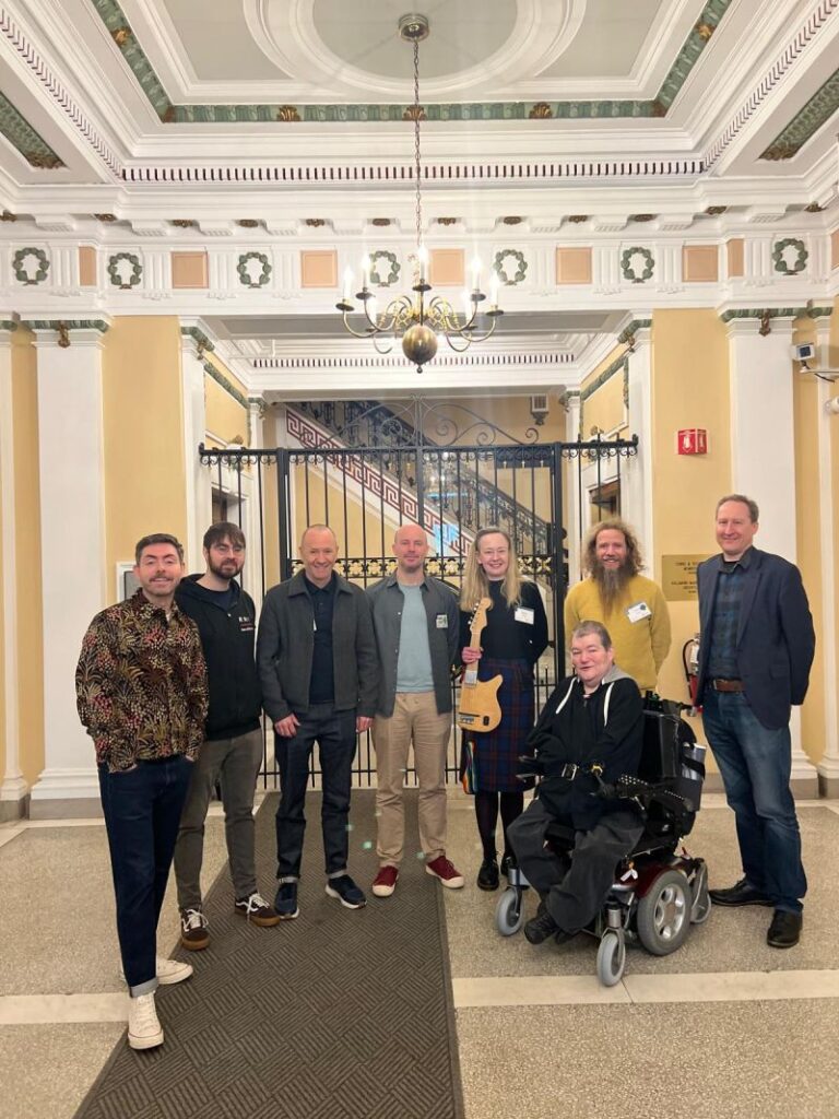 Eight people pose for a group photo in an elegant building with ornate ceilings and a chandelier. One person sits in a motorized wheelchair at the front; the others stand behind, smiling. A staircase and decorative railings are in the background.