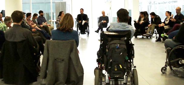 A group of people, some using wheelchairs, sit in a circle in a bright, modern room, engaged in a discussion or meeting.