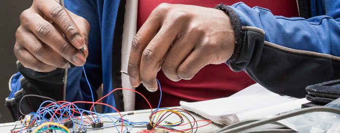 A person connects colorful wires on an electronic circuit board, working with their hands. They are wearing a blue jacket and a red shirt.