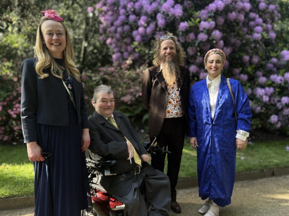 Four people pose together outdoors in front of purple flowering bushes. One person is in a wheelchair, and the others stand around them, all dressed in formal or semi-formal attire, smiling at the camera.