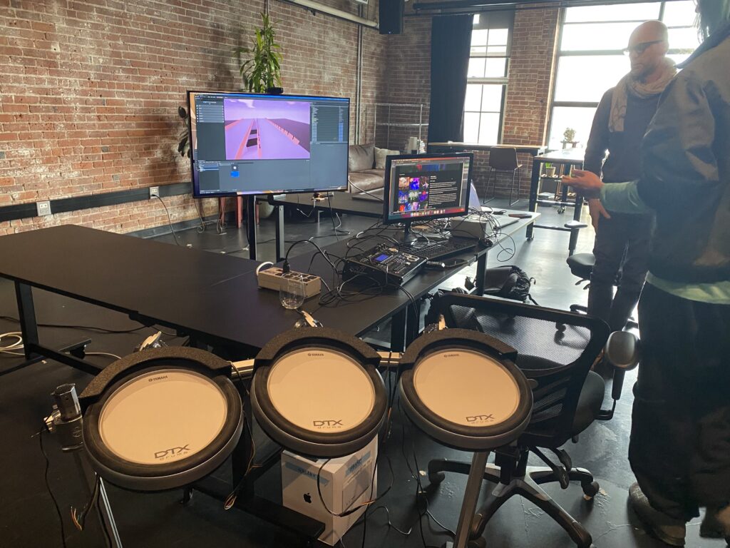 Four electronic drum pads are set up in front of a table with computer monitors, cables, and electronics in an industrial-style room. Two people stand nearby, and a large screen displays a digital interface.