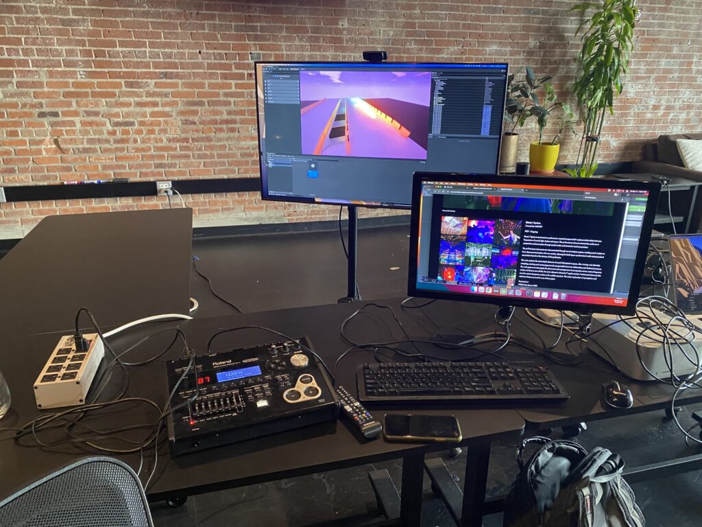 A workspace with two monitors on a desk, one displaying a 3D graphics program and the other showing a colorful website. Audio equipment, a keyboard, mouse, and other electronics are also on the desk. Brick wall and plants in background.
