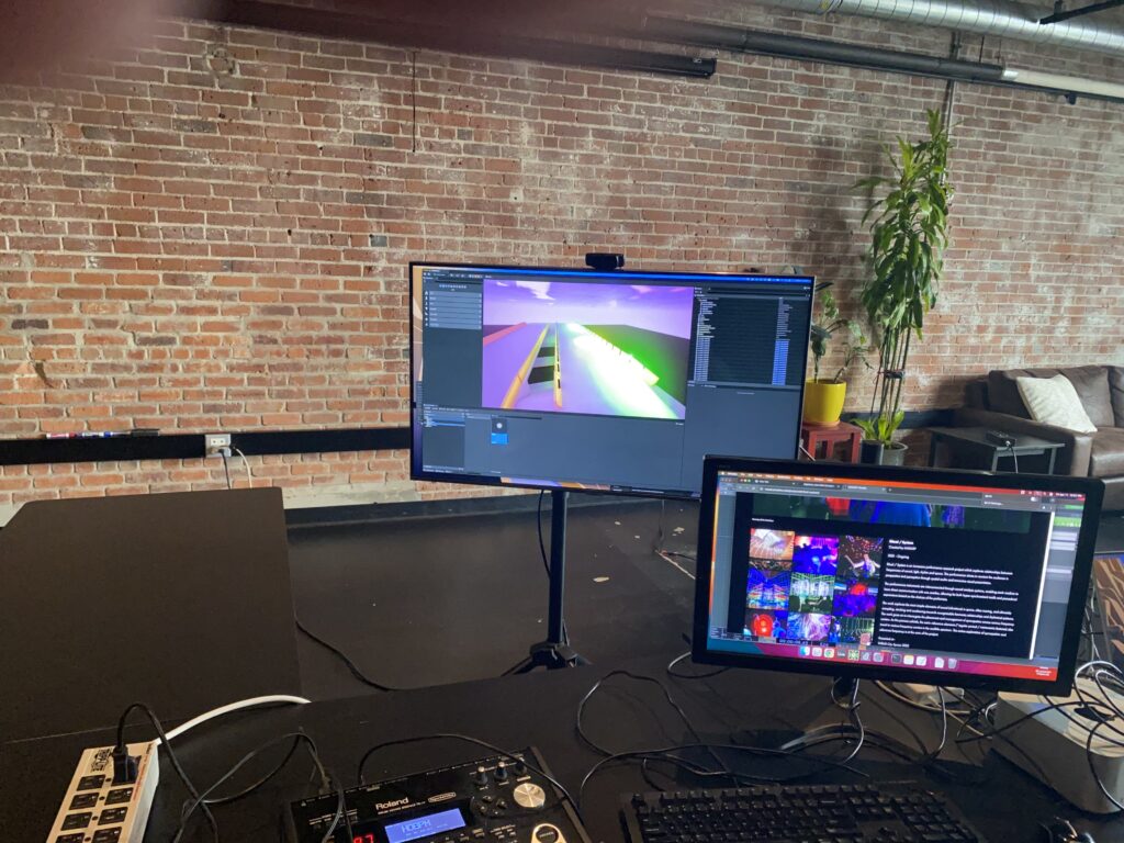 Two computer monitors on a desk in a loft-style room with an exposed brick wall; one screen displays a colorful 3D road scene, and the other shows a webpage. Plants and a couch are in the background.