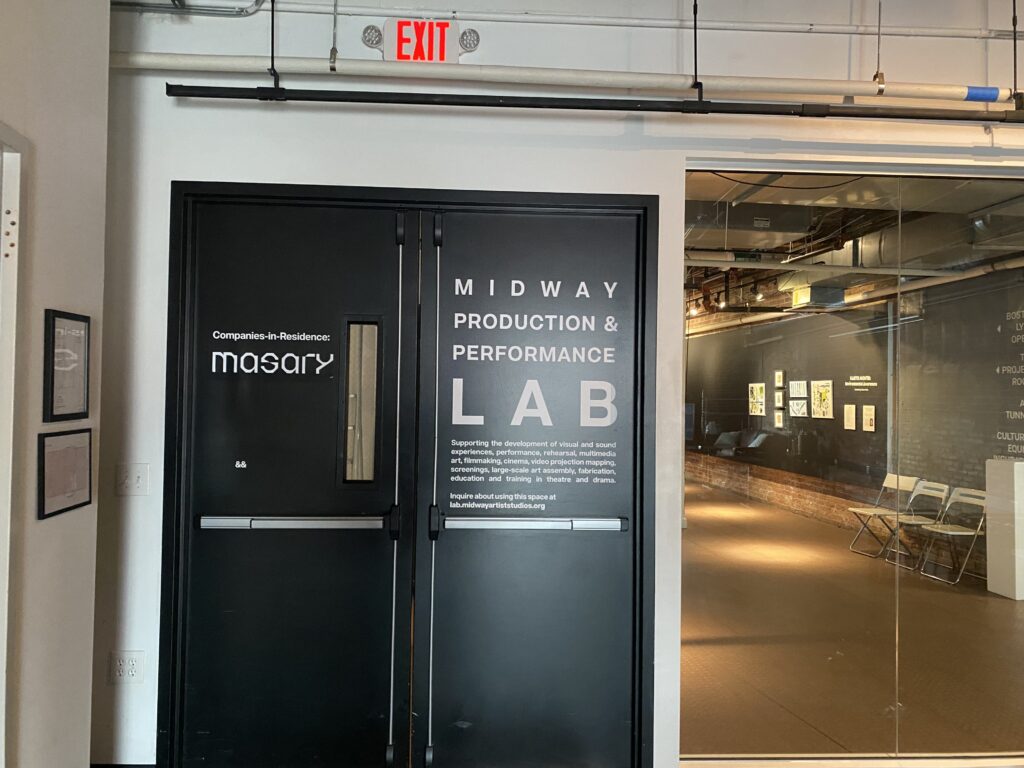 Double black doors labeled Midway Production & Performance Lab next to a glass wall showing an interior space with artworks displayed on brick and white walls. An exit sign hangs above the doors.