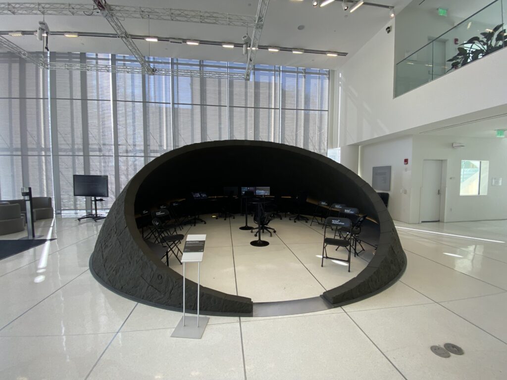 A large, dome-shaped black structure with open entrance sits in a bright, modern lobby with glass walls; inside are several folding chairs and computer monitors arranged in a semicircle.