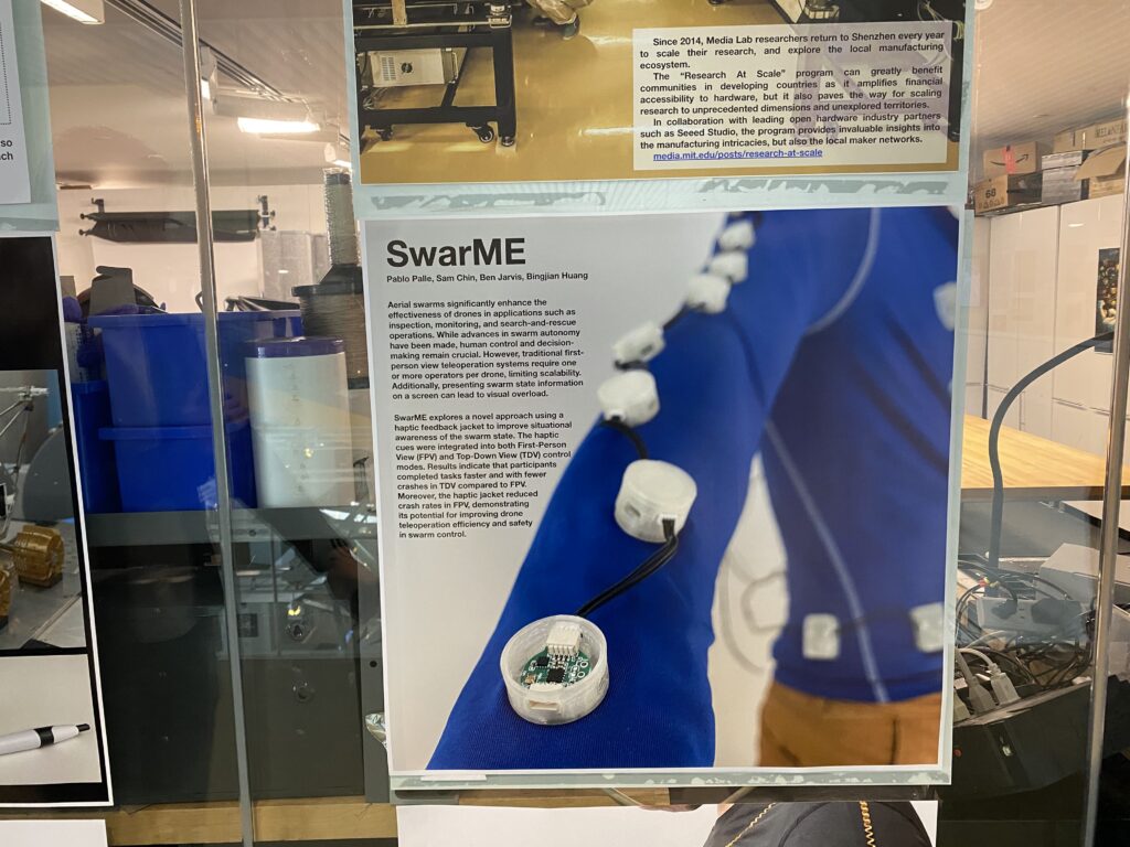 A display shows a poster titled SwarME with a photo of a persons arm wearing a blue sleeve fitted with white circular sensor devices. A close-up reveals an open sensor exposing electronic components.