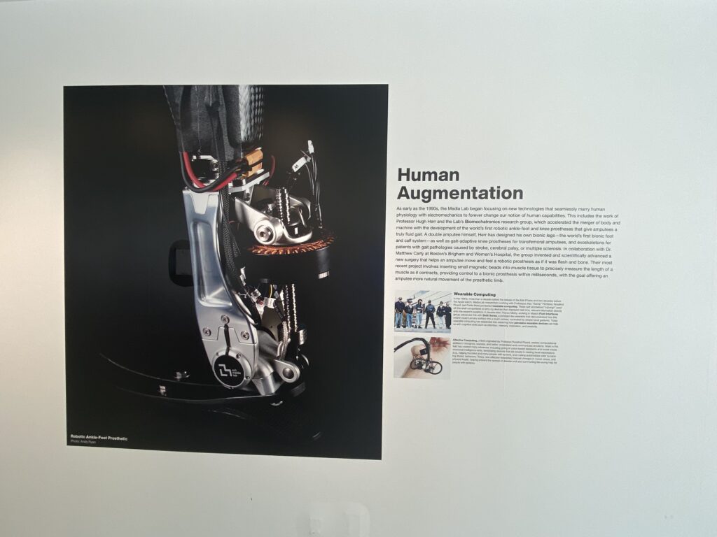 Photo of a museum display titled Human Augmentation featuring a close-up image of a prosthetic running blade with explanatory text and smaller photos showing athletes and wearable technology.