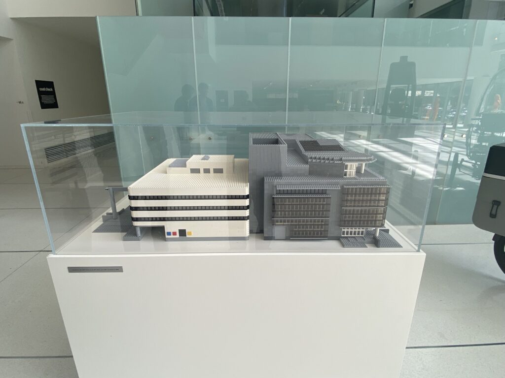 A scale model of two modern buildings, constructed with LEGO bricks, is displayed in a clear glass case on a white pedestal in a bright, contemporary indoor space.