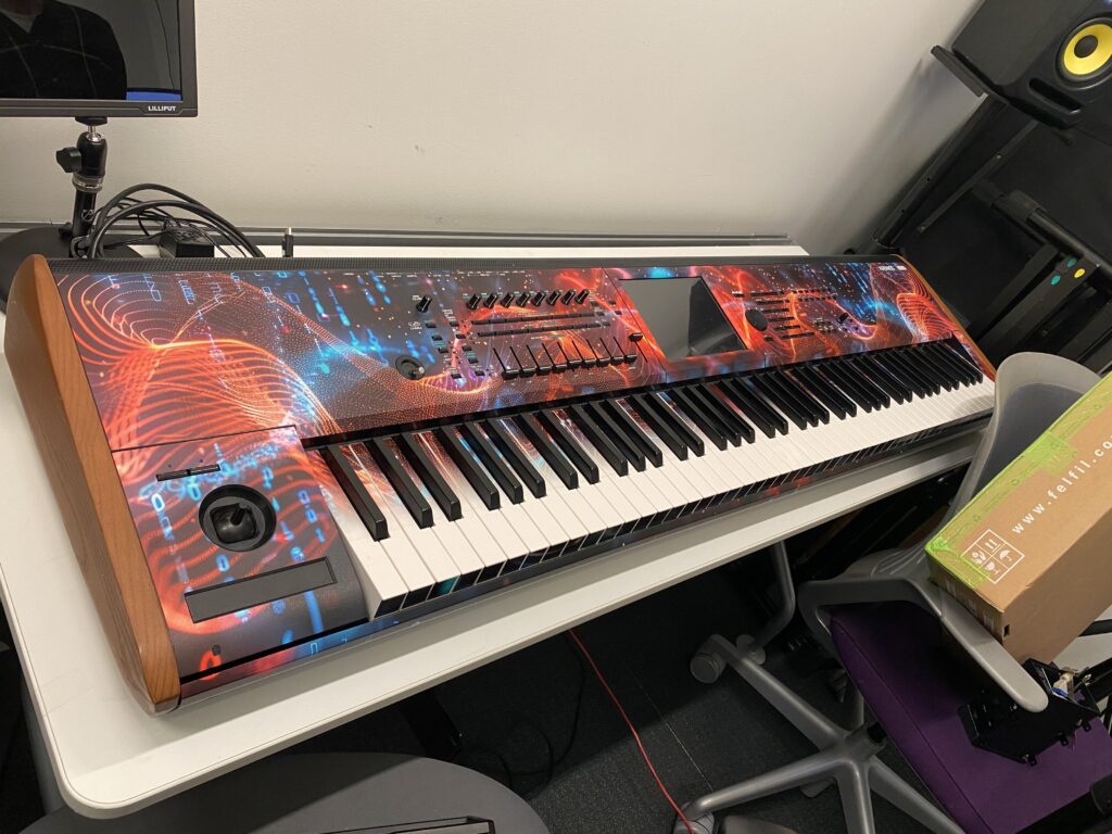 A colorful synthesizer keyboard with a vibrant, abstract red and blue design sits on a white table next to a cardboard box and various electronic equipment.