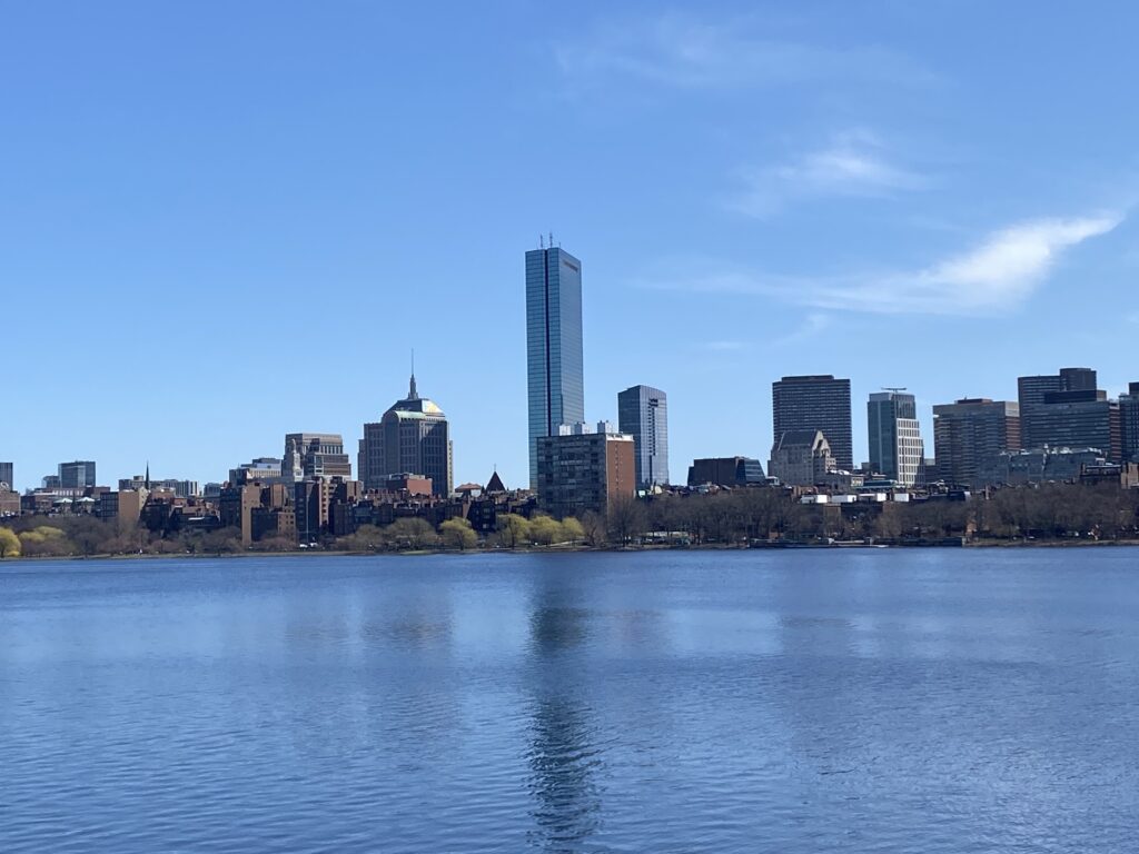 A city skyline with modern and historic buildings stands under a blue sky, reflected in the calm water of a river in the foreground, with some green trees lining the riverbank.