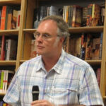 A man wearing glasses and a plaid short-sleeve shirt holds a microphone while standing in front of bookshelves filled with books.