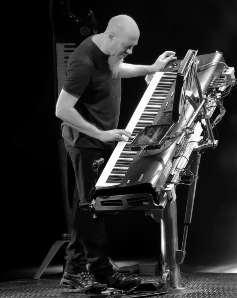 A man with a shaved head and beard plays an electronic keyboard on stage, surrounded by multiple synthesizers and music equipment, illuminated by stage lights in a dark setting.