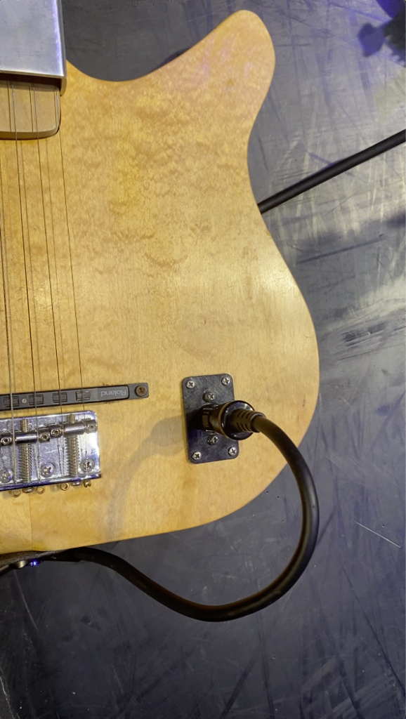 Close-up of a natural wood electric guitar body, showing the bridge, strings, and a black cable plugged into the input jack on a dark, textured surface.