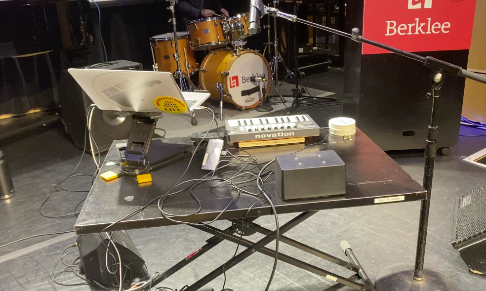 A music setup on a stage includes a laptop, Novation MIDI controller, cables, and a small audio device on a black table. A drum set and a red Berklee sign are visible in the background.