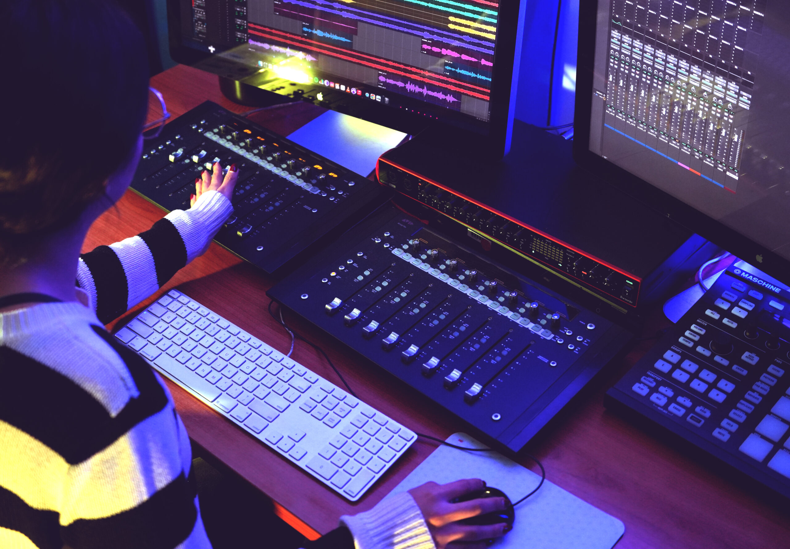 A person working at a music production studio desk with audio mixing equipment, a computer keyboard, and multiple monitors displaying colorful audio tracks and mixing software.