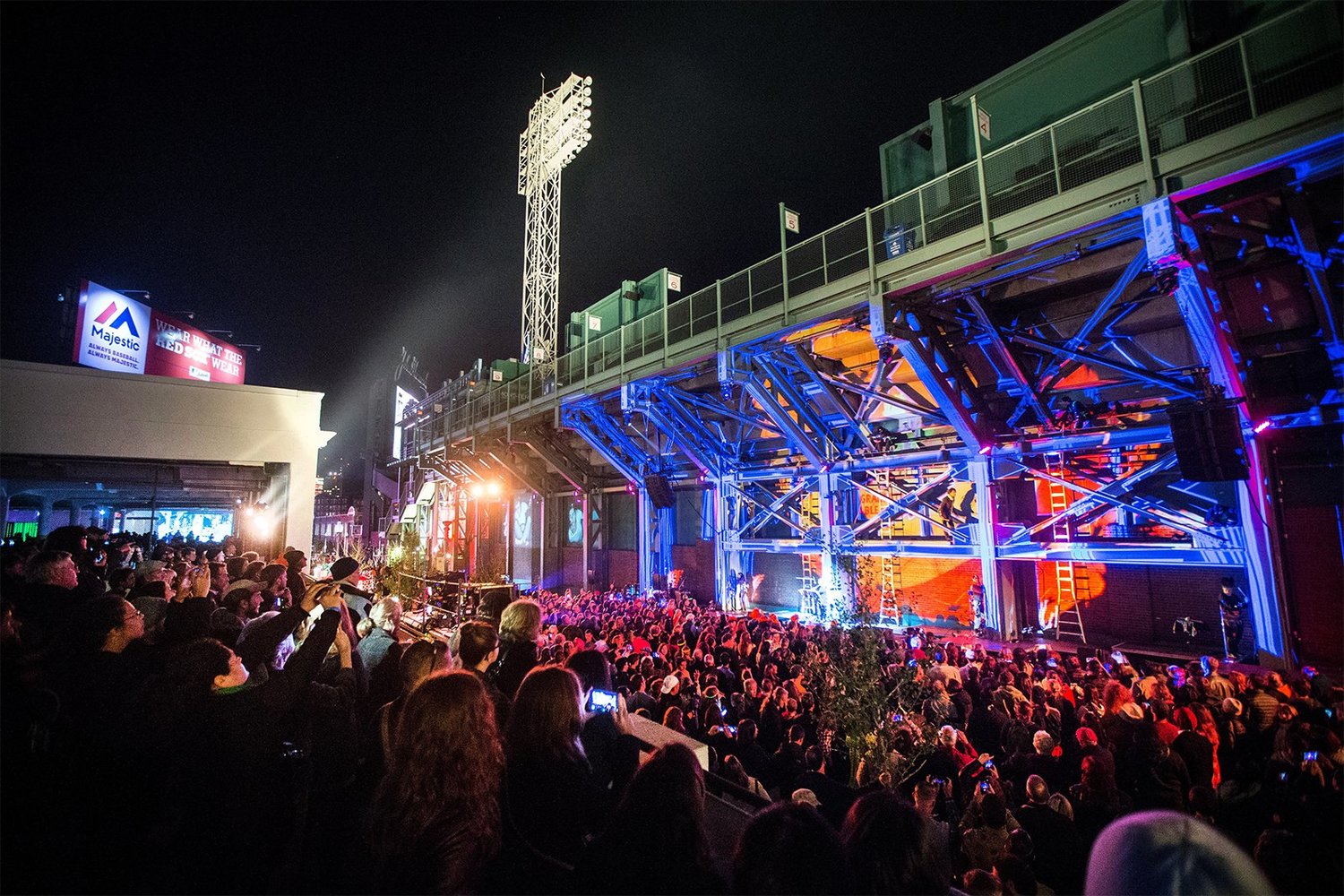 A large crowd gathers at night under stadium lights and metal beams, watching a live performance with colorful stage lighting at an outdoor venue.