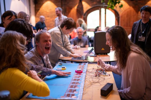 People gather around a table with electronic components and wires. A man laughs energetically while others interact with the setup in a bright, lively room with large windows and brick walls.