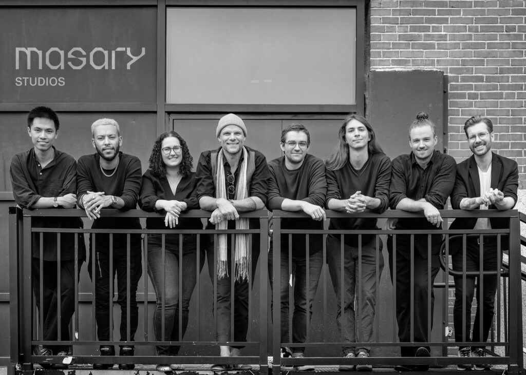 A picture of the members of eight members if Masary Studios leaning against a fence in front of the building their offices are in