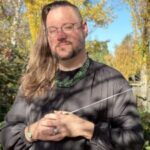 A person with long hair, glasses, and a beard stands outdoors in sunlight, wearing a dark top and holding a conductor’s baton. Autumn trees with yellow leaves are in the background.