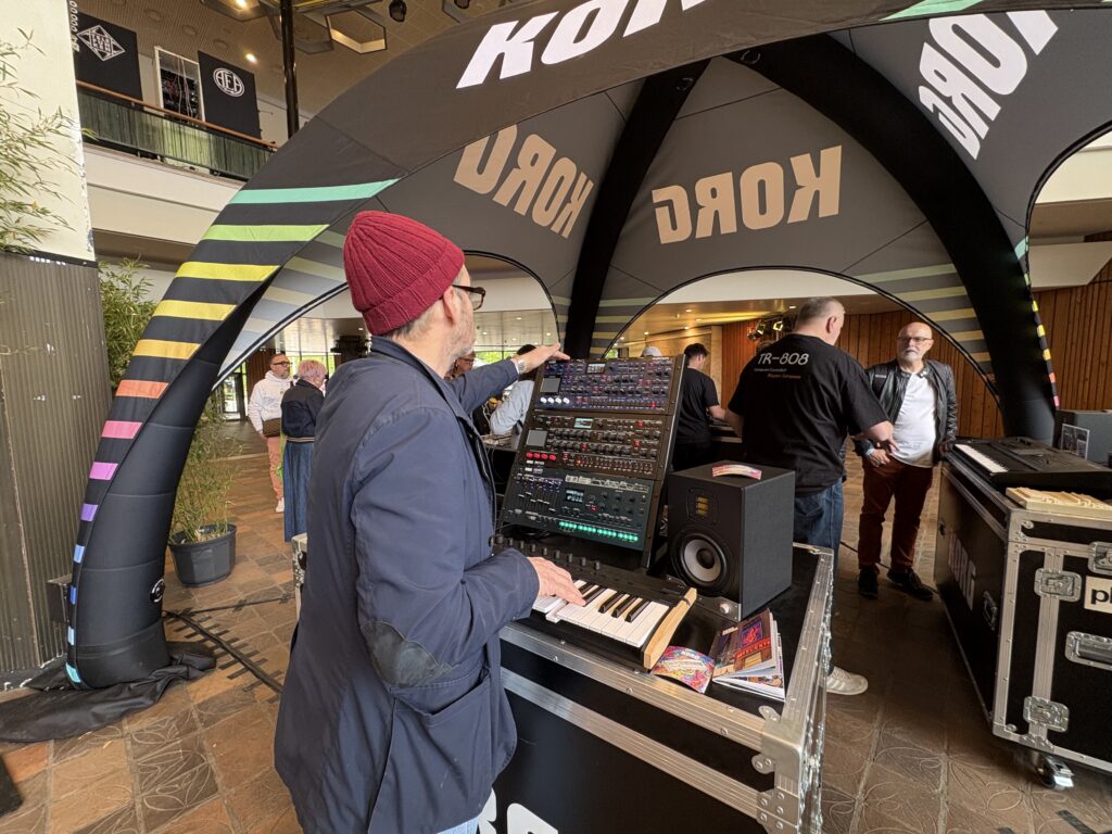 A man wearing a red beanie and glasses plays a synthesizer at a Korg booth under a branded tent. Several people stand nearby, and the setting appears to be an indoor event or exhibition.
