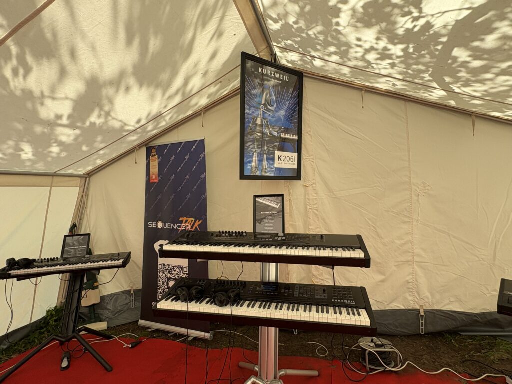 Two electronic keyboards are displayed on a stand inside a tent, with a red carpet underneath. A poster about synthesizers labeled K2001 and banners are in the background, along with another keyboard on a stand.