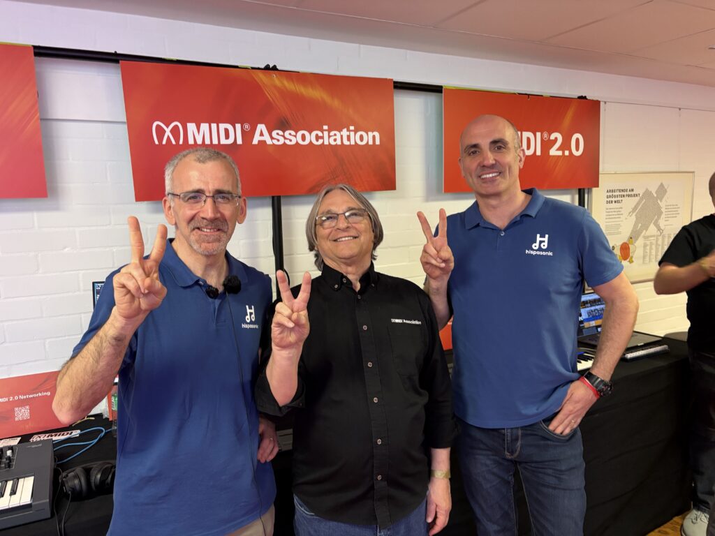 Three smiling people stand indoors in front of red MIDI Association signs, each holding up a peace sign with their fingers. The two on the left and right wear matching blue polo shirts, and the center person wears a black shirt.