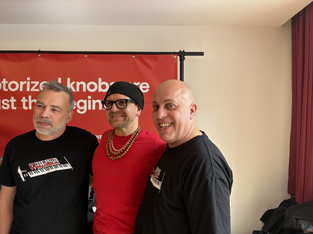 Three smiling men stand together indoors in front of a red banner with white text. Two wear black t-shirts and one wears a red shirt, glasses, a black beanie, and a beaded necklace.