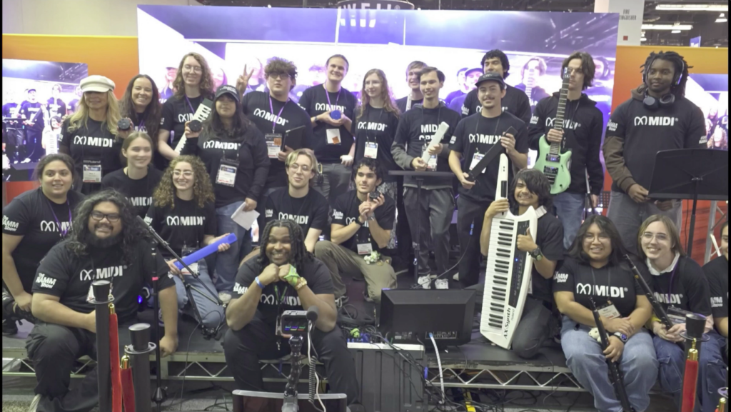 A large group of young people wearing matching black “MIDI” shirts pose together on a stage with musical instruments, including guitars and a keytar. Everyone is smiling and looking at the camera.