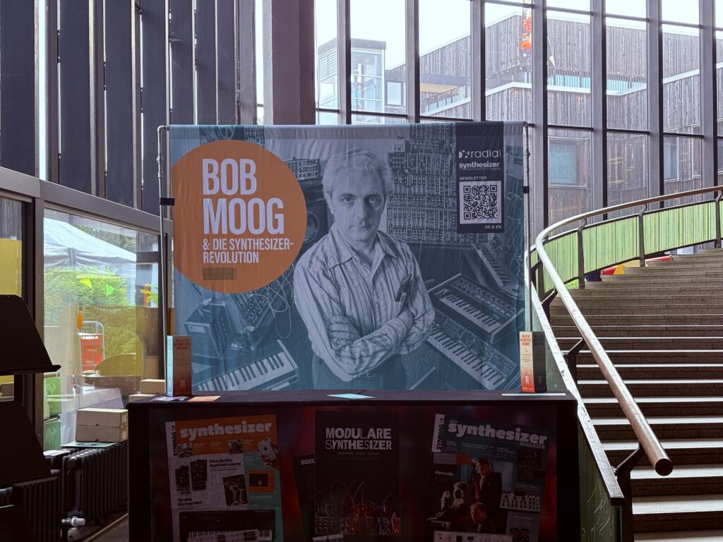 A display featuring a large poster of Bob Moog with synthesizers, text reading Bob Moog & Die Synthesizer-Revolution, and magazines titled Synthesizer on a table in a modern, glass-walled building.