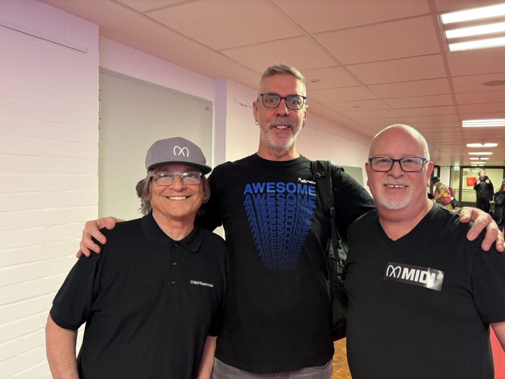 Three smiling men stand together indoors under pink lighting. The man on the left wears a gray cap and black polo shirt, the middle man wears glasses and an AWESOME t-shirt, and the right man wears glasses and a MIDI t-shirt.