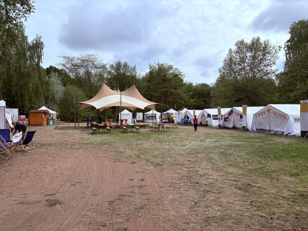 A grassy outdoor area with several white tents and a shaded canopy structure at the center, surrounded by trees. A few people and empty chairs are visible, creating a campsite or festival atmosphere.