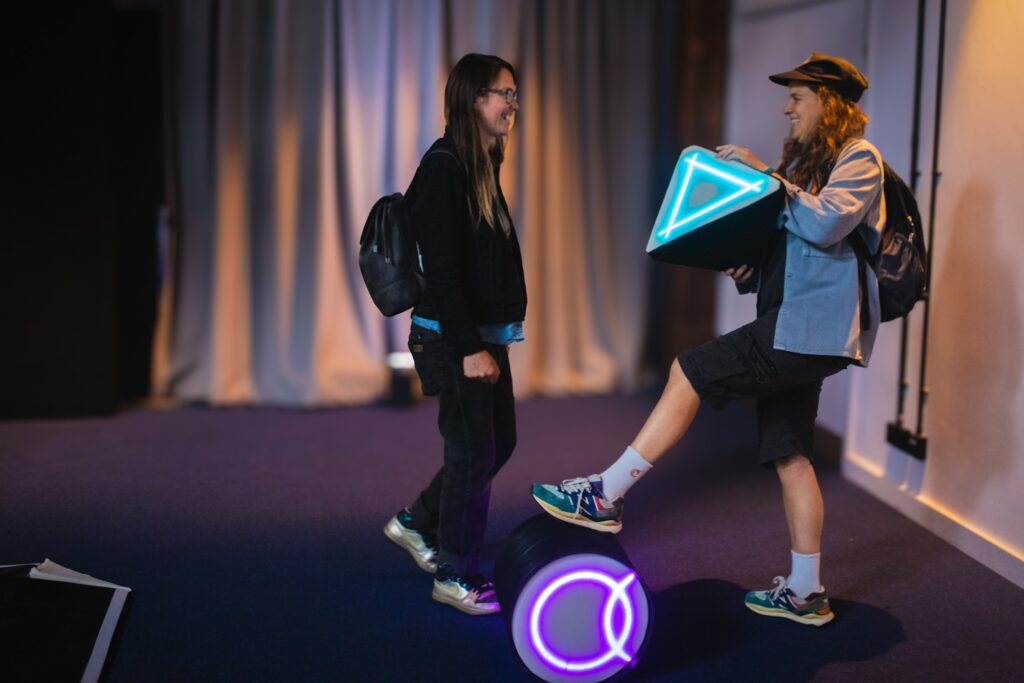 Two people with backpacks stand indoors; one balances a glowing triangular object while stepping on a round object with a neon symbol. The room is softly lit with curtains in the background.